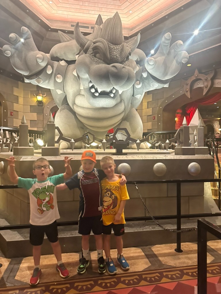 Three boys excitedly posing in front of a giant Bowser statue in the Mario Kart: Bowser's Challenge queue at Epic Universe