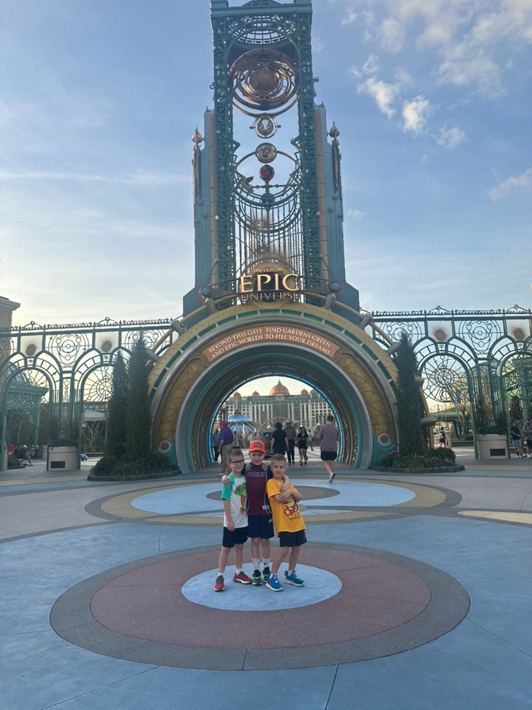 The ornate Epic Universe entrance gate and clock tower at dusk, with three boys standing beneath the archway in the distance